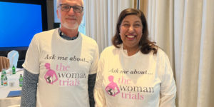 Photograph of a man and woman smiling at the camera wearing t-shirts that say "Ask me about the WOMAN Trials"